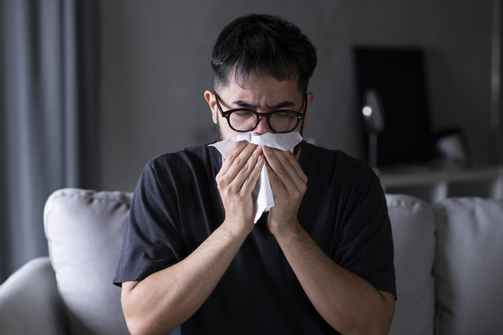 Asian man sick and sneeze with tissue paper on sofa at home. Male blowing nose suffering from cold. Cold and fever concept.